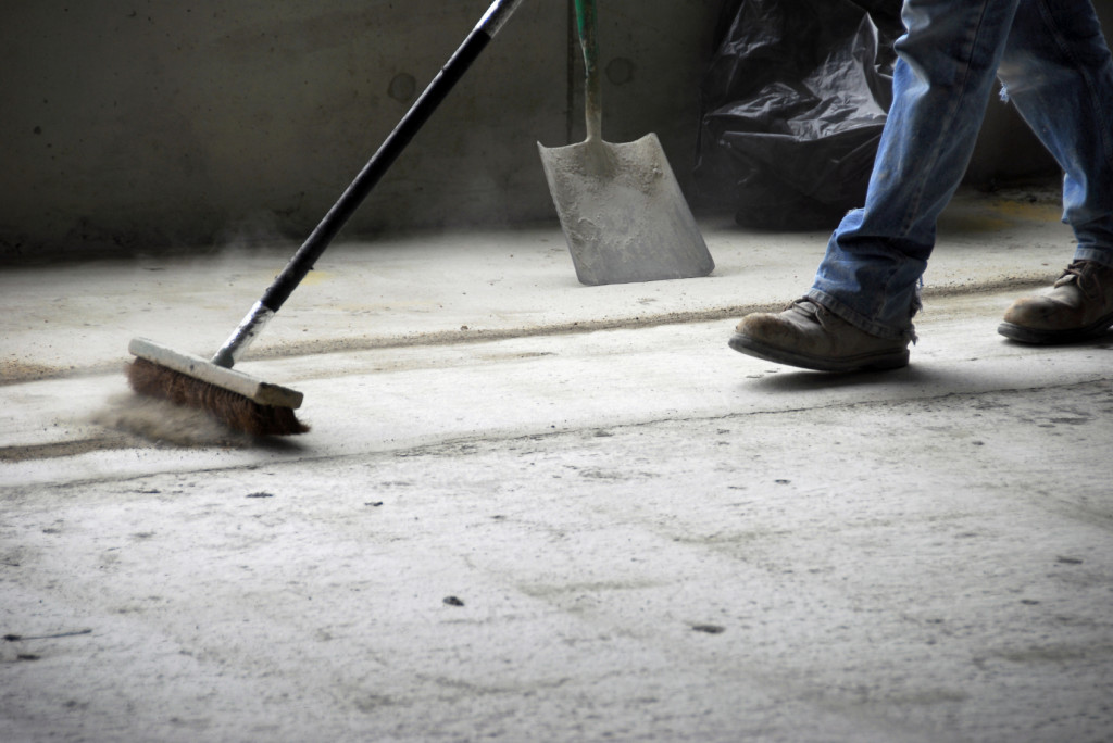 The legs and feet of a construction worker sweeping up on rough concrete at a job site using a large broom